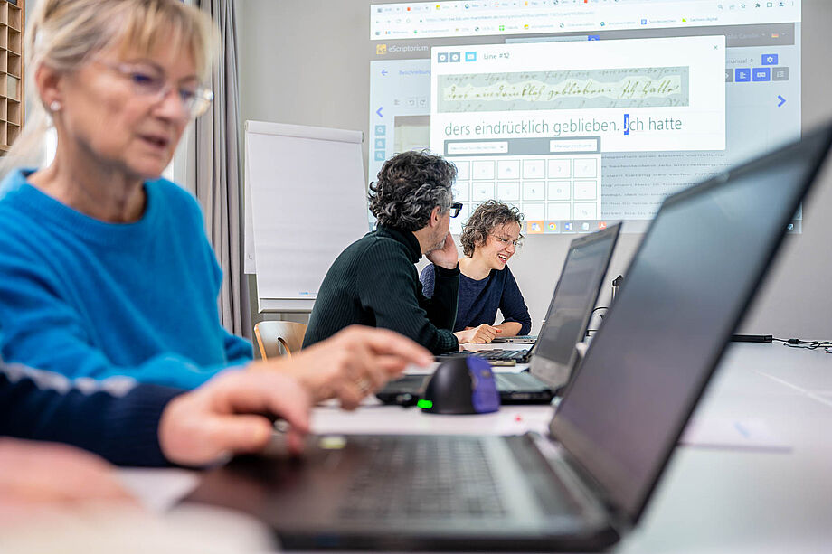 Drei Personen besprechen sich am Tisch vor PC-Bildschirm, Handschrift an der Leinwand im Hintergrund